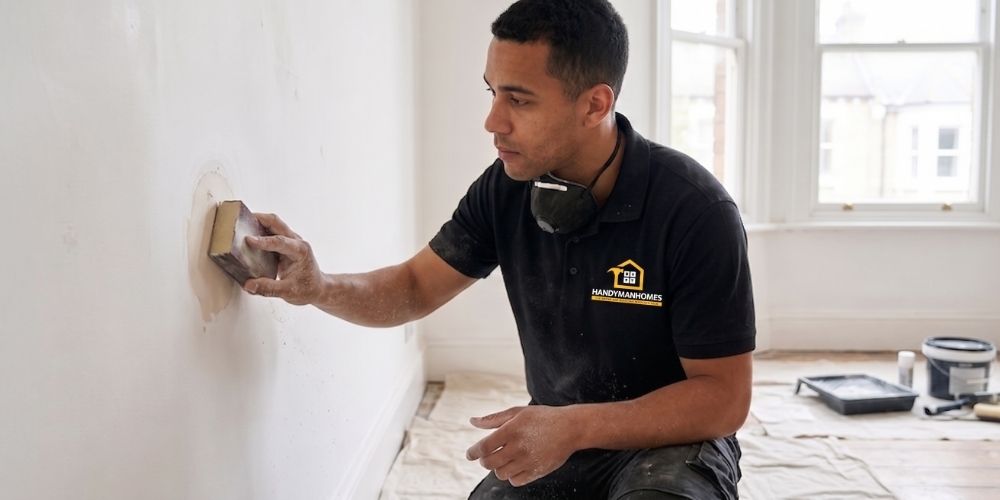 A painter using a sanding block to level a dried wall patch, with drop sheets protecting the timber floors.
